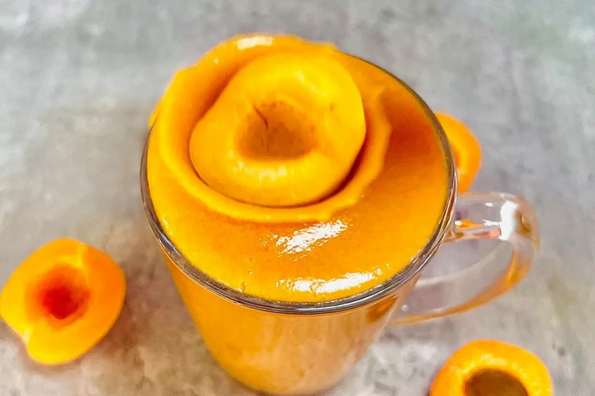 a slice of apricot being dropped onto a glass cup filled with an apricot smoothie