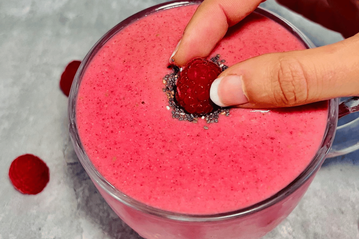 a hand placing a fresh raspberry on top of a smoothie cup