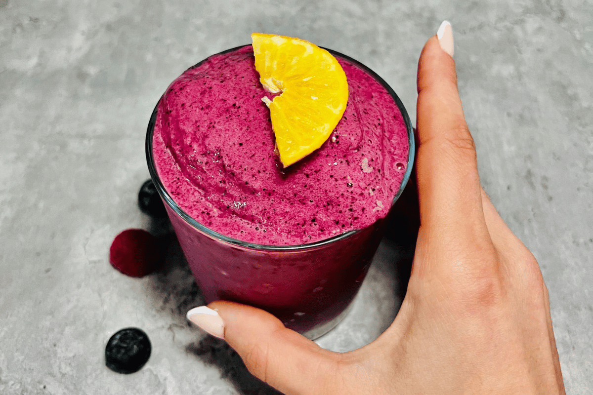 a hand holding a glass cup filled with berry smoothie topped with a slice of orange