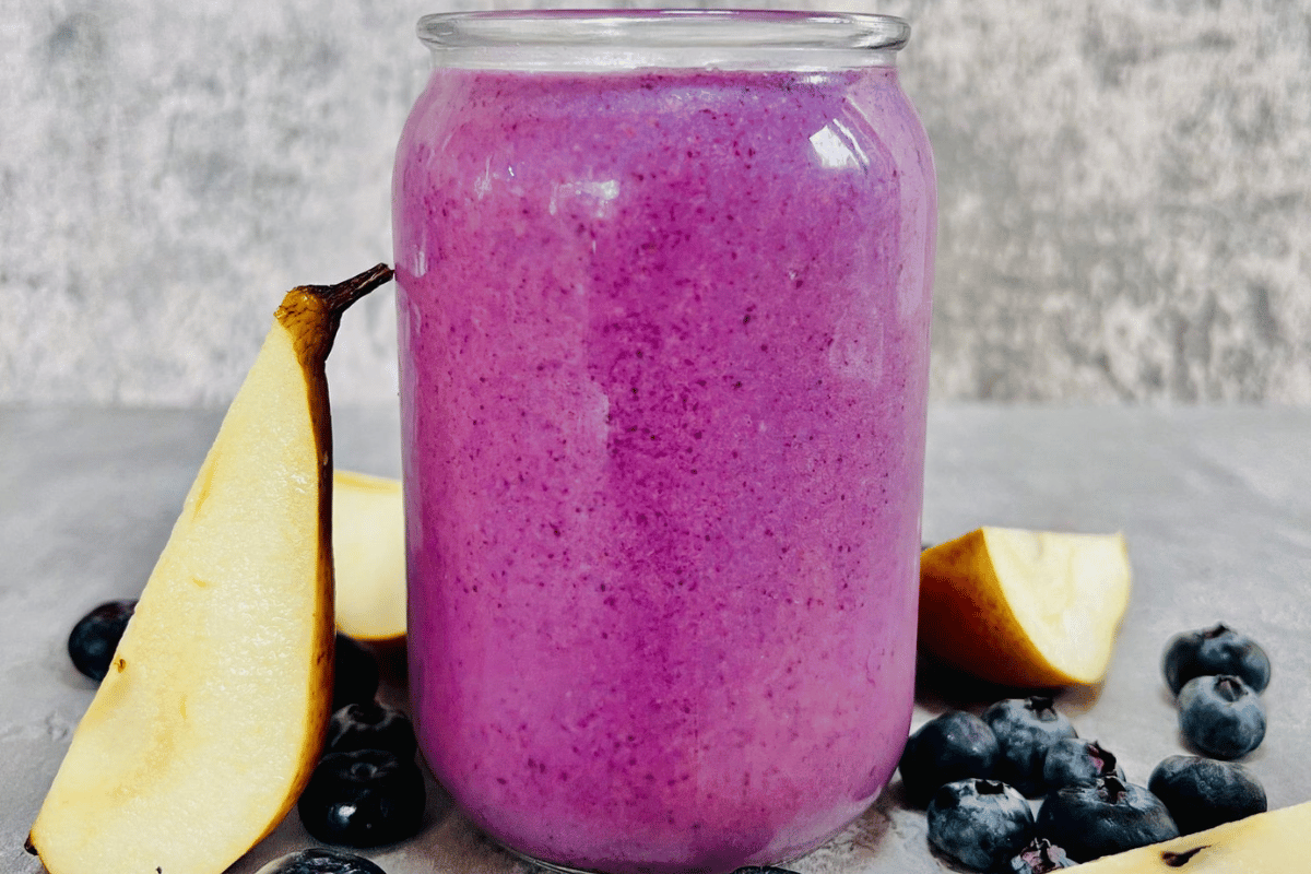 blueberry smoothie in a glass cup surrounded by fresh blueberries and a sliced pear