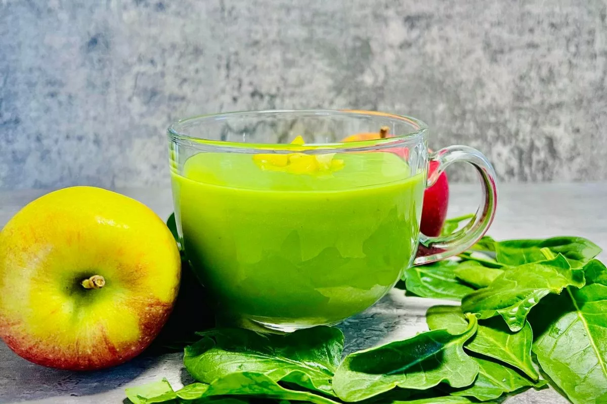 a glass cup filled with a green smoothie, surrounded by an apple and spinach