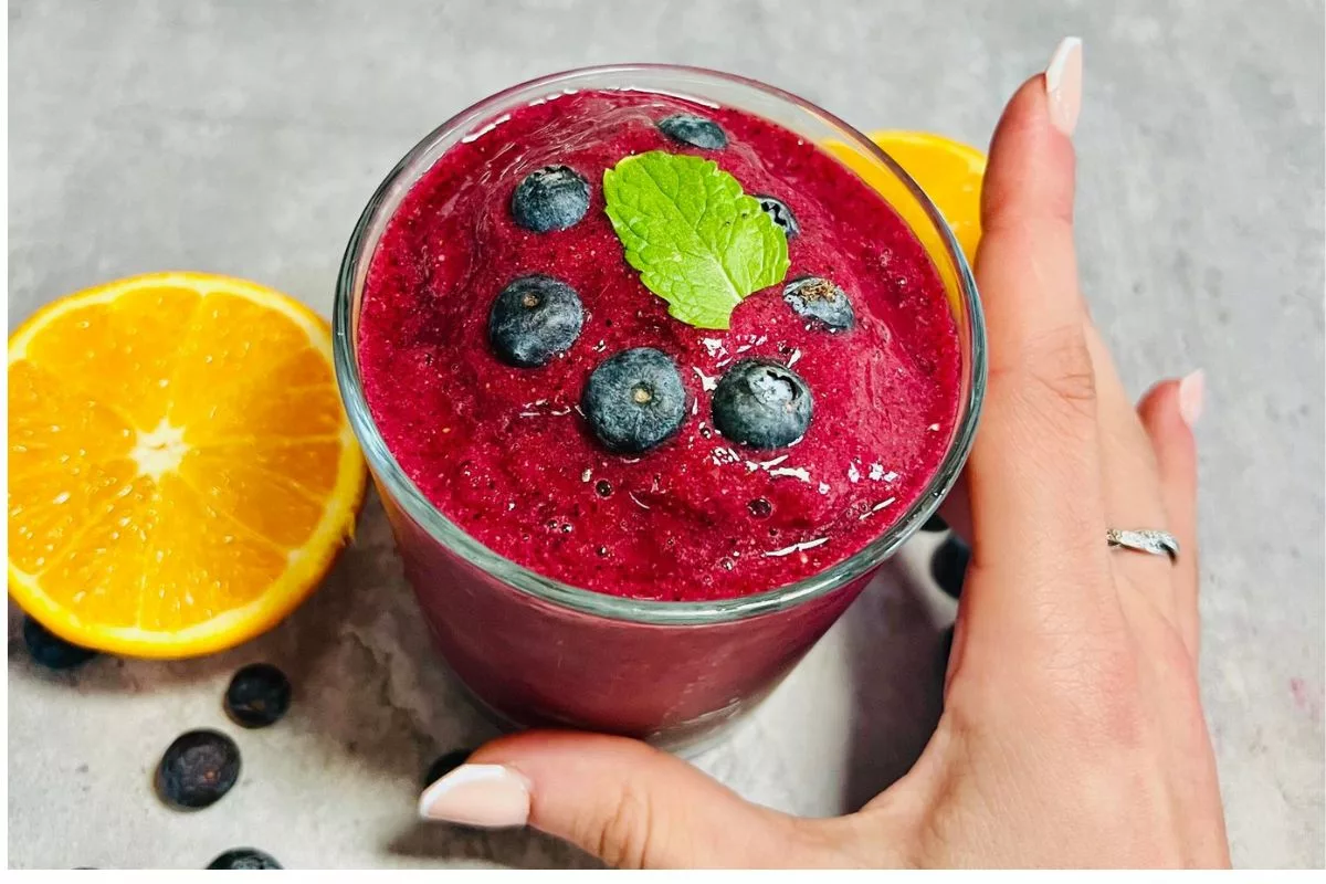 a hand holding a glass cup filled with berry smoothie topped with fresh mint and blueberries