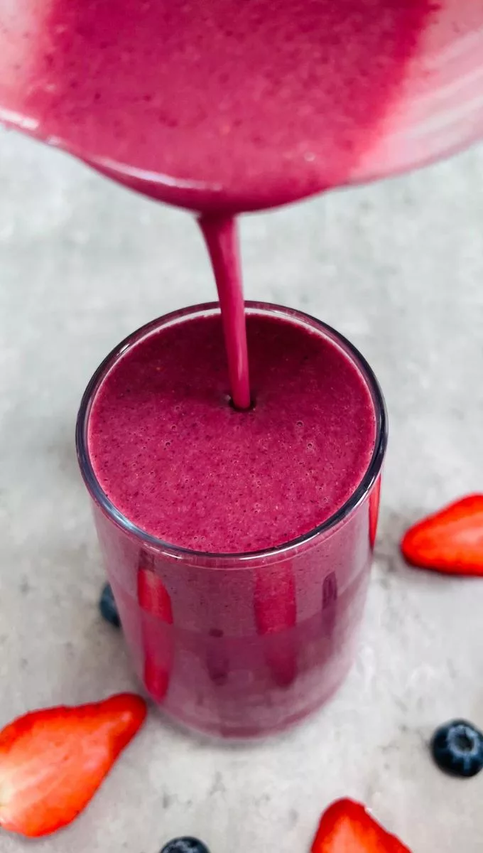 Strawberry Banana Blueberry Smoothie being poured into a tall thin glass cup from a blender jug