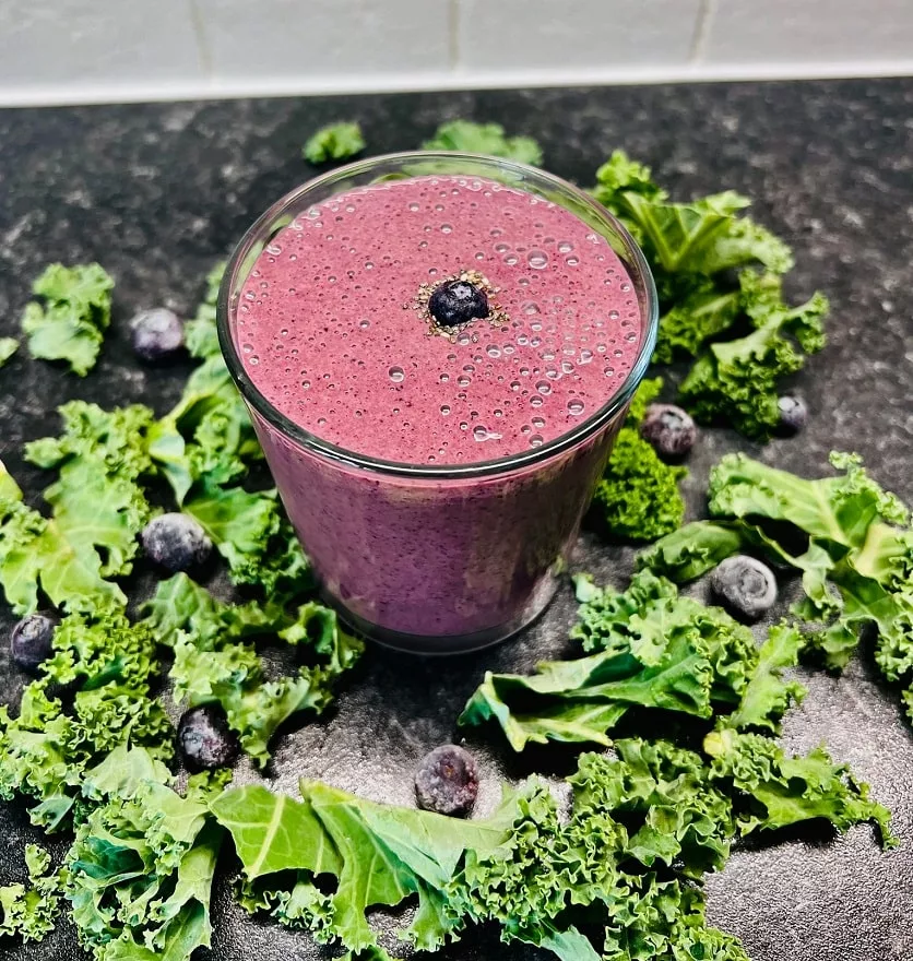 blueberry smoothie made with kale and kefir, served in a glass cup surrounded by kale and blueberries