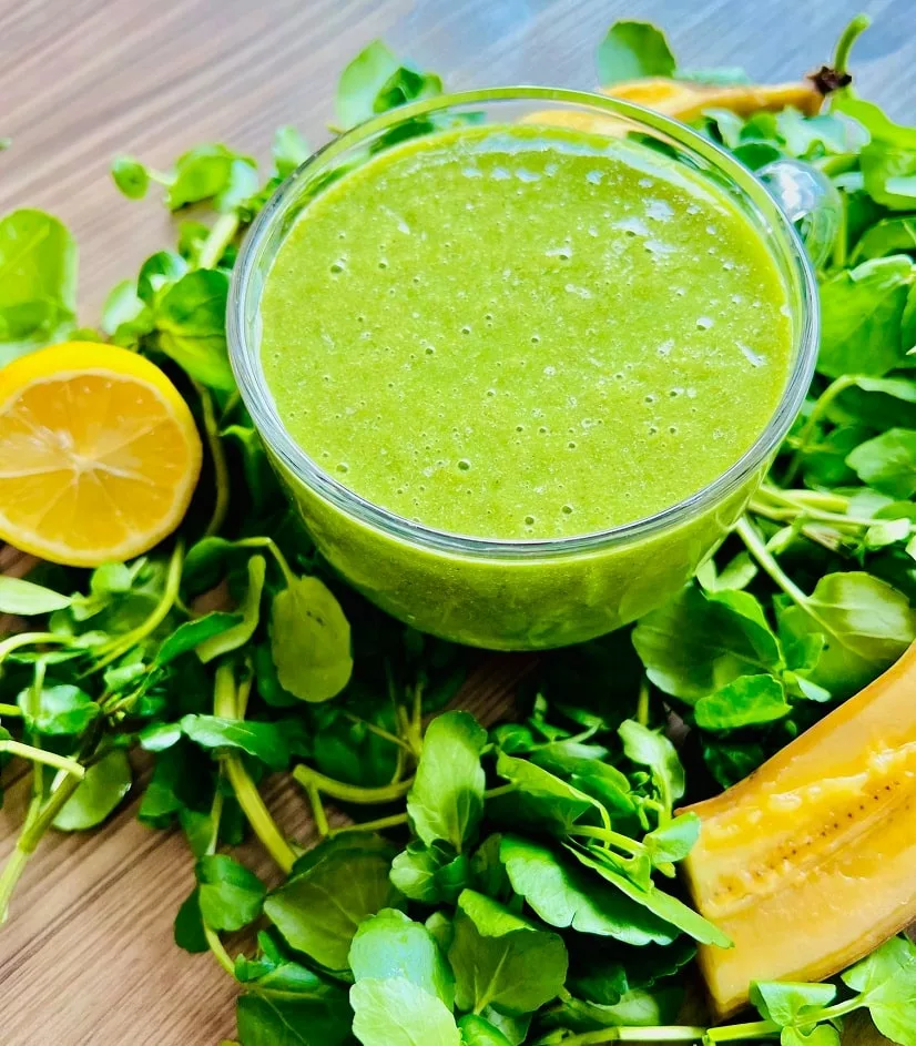 Watercress Smoothie surrounded by fresh watercress, lemon, and sliced banana on a wooden surface