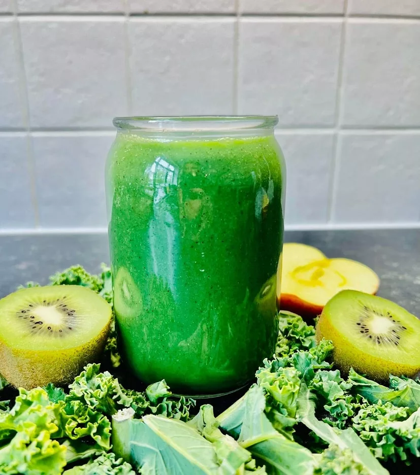 green smoothie served in a tall glass cup surrounded by fresh kale, sliced kiwi and apple