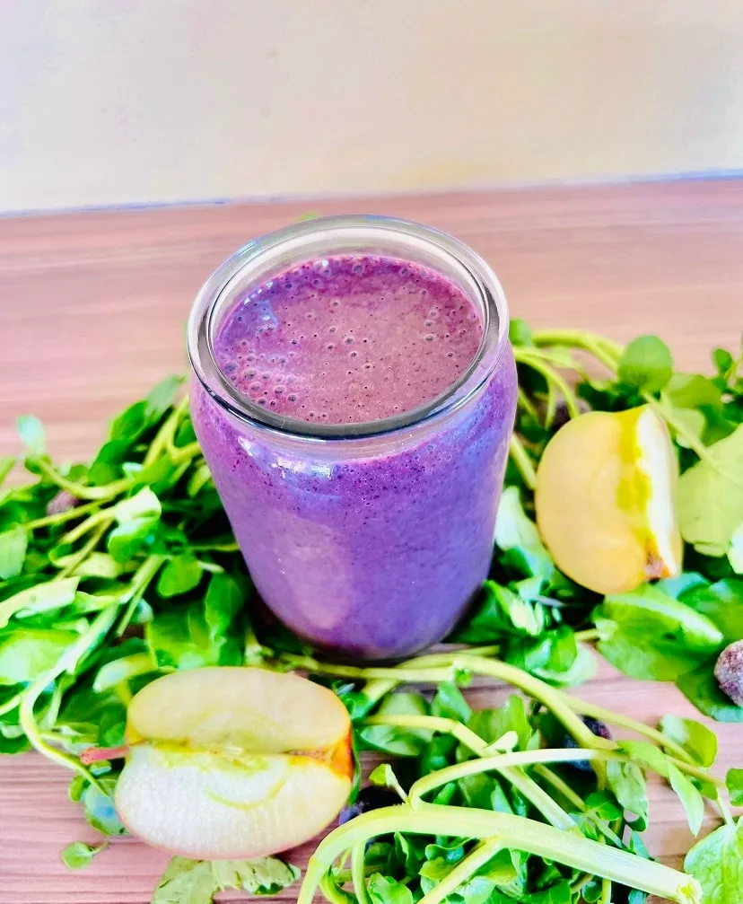 Watercress Blueberry Smoothie served in a glass cup, surrounded by sliced apple, blueberries and watercress on a wooden surface. 