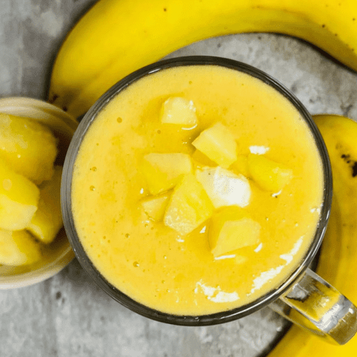 pineapple smoothie served in a tall glass cup surrounded by two bananas and a cup of frozen pineapple slices