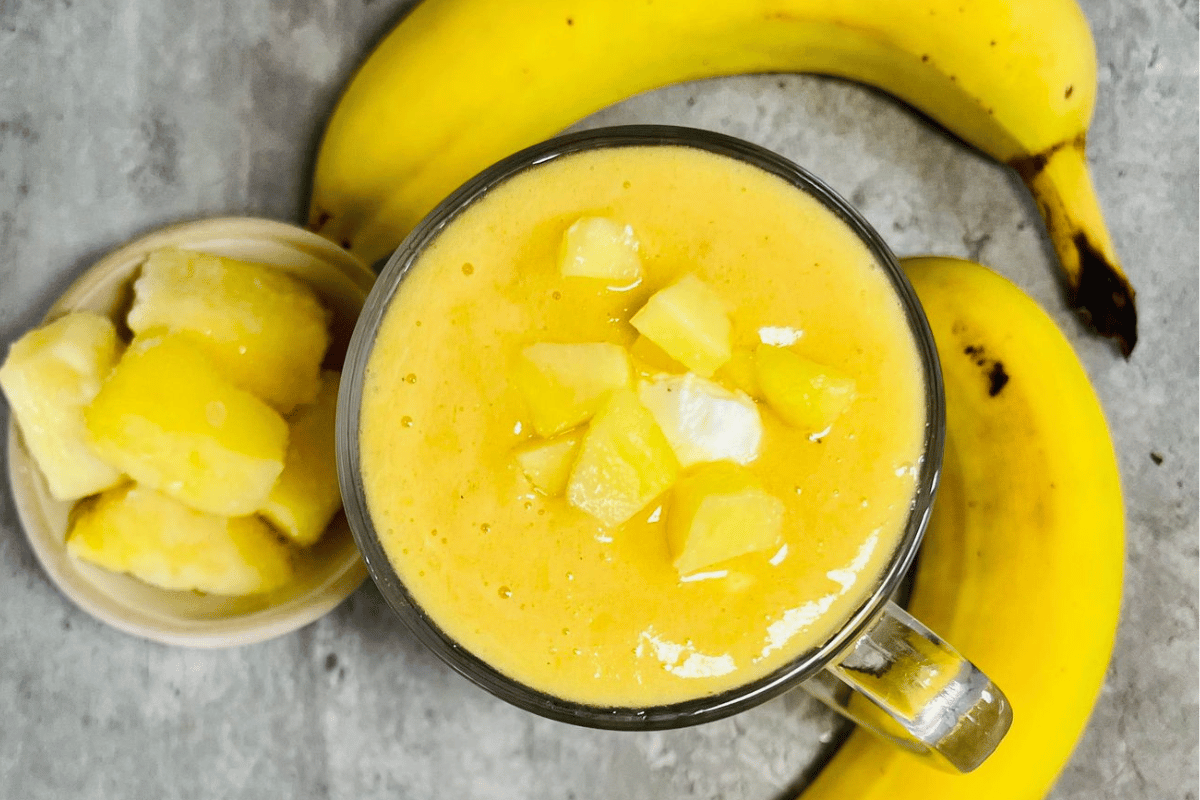 pineapple smoothie served in a tall glass cup surrounded by two bananas and a cup of frozen pineapple slices