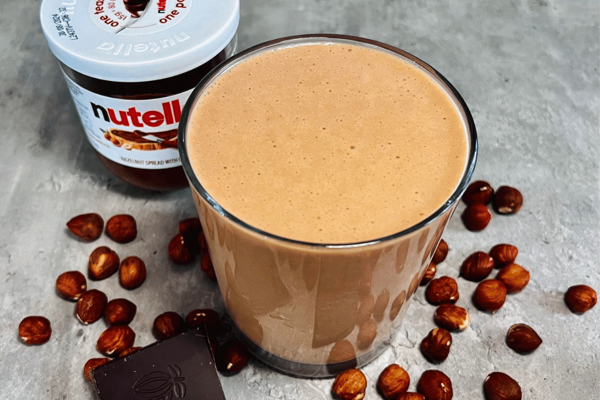 a brown smoothie served in a glass cup next to a nutella jar surrounded by hazelnuts