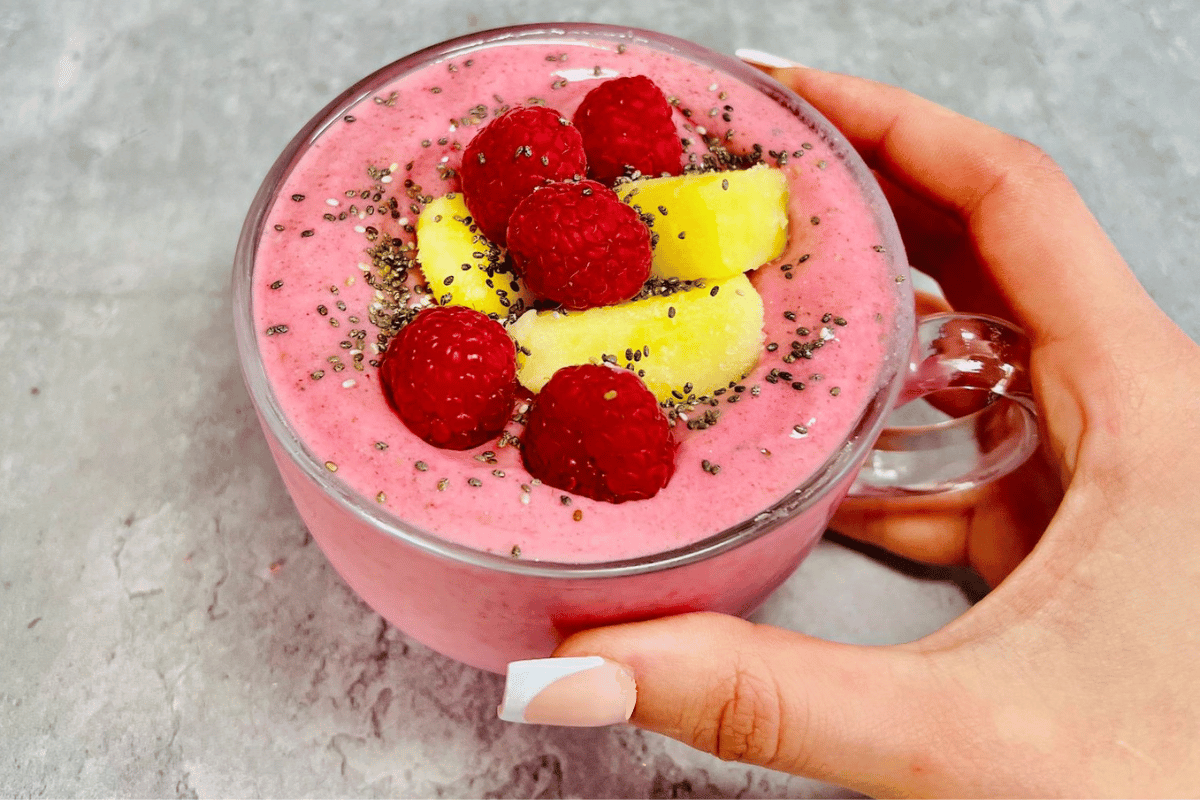 a hand holding a glass cup filled with pineapple and a raspberry smoothie