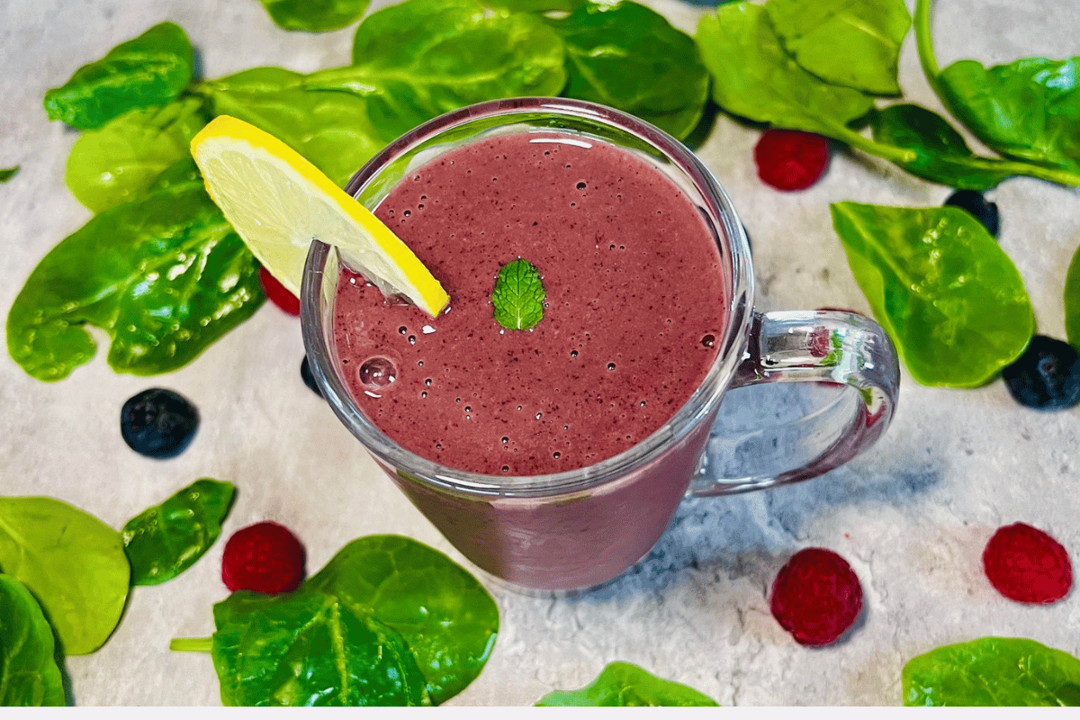 blueberry smoothie filled glass surrounded by spinach and a slice of lemon on top of it