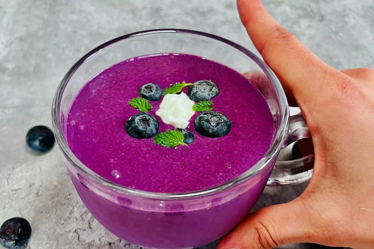 a hand holding a round glass cup of blueberry smoothie