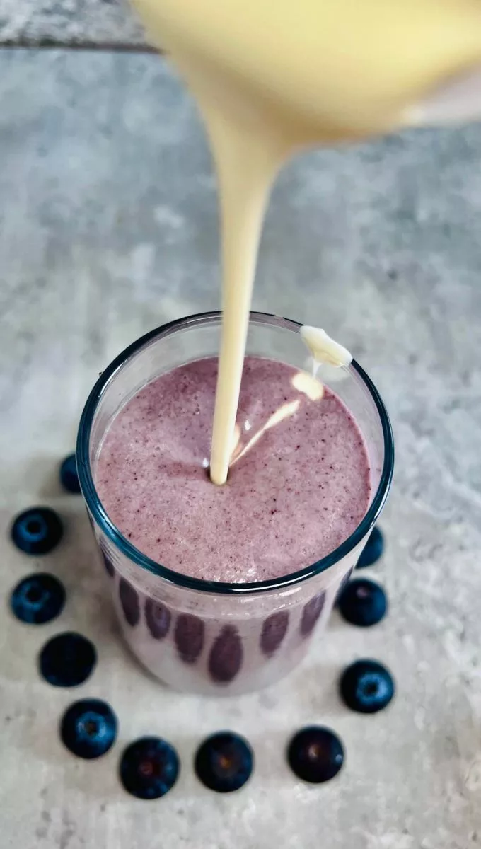 heavy cream being poured into a glass cup smoothie filled with blended blueberry smoothie