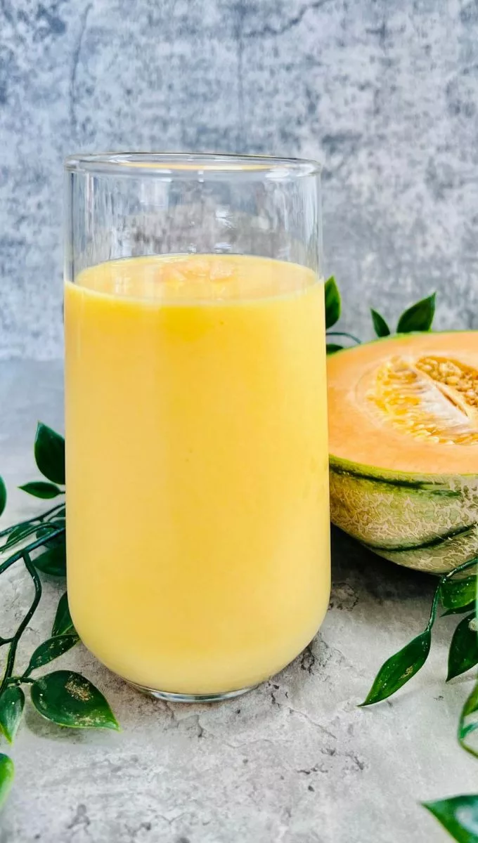 Cantaloupe Smoothie served in a tall thin glass cup surrounded by greens and a fresh cantaloupe